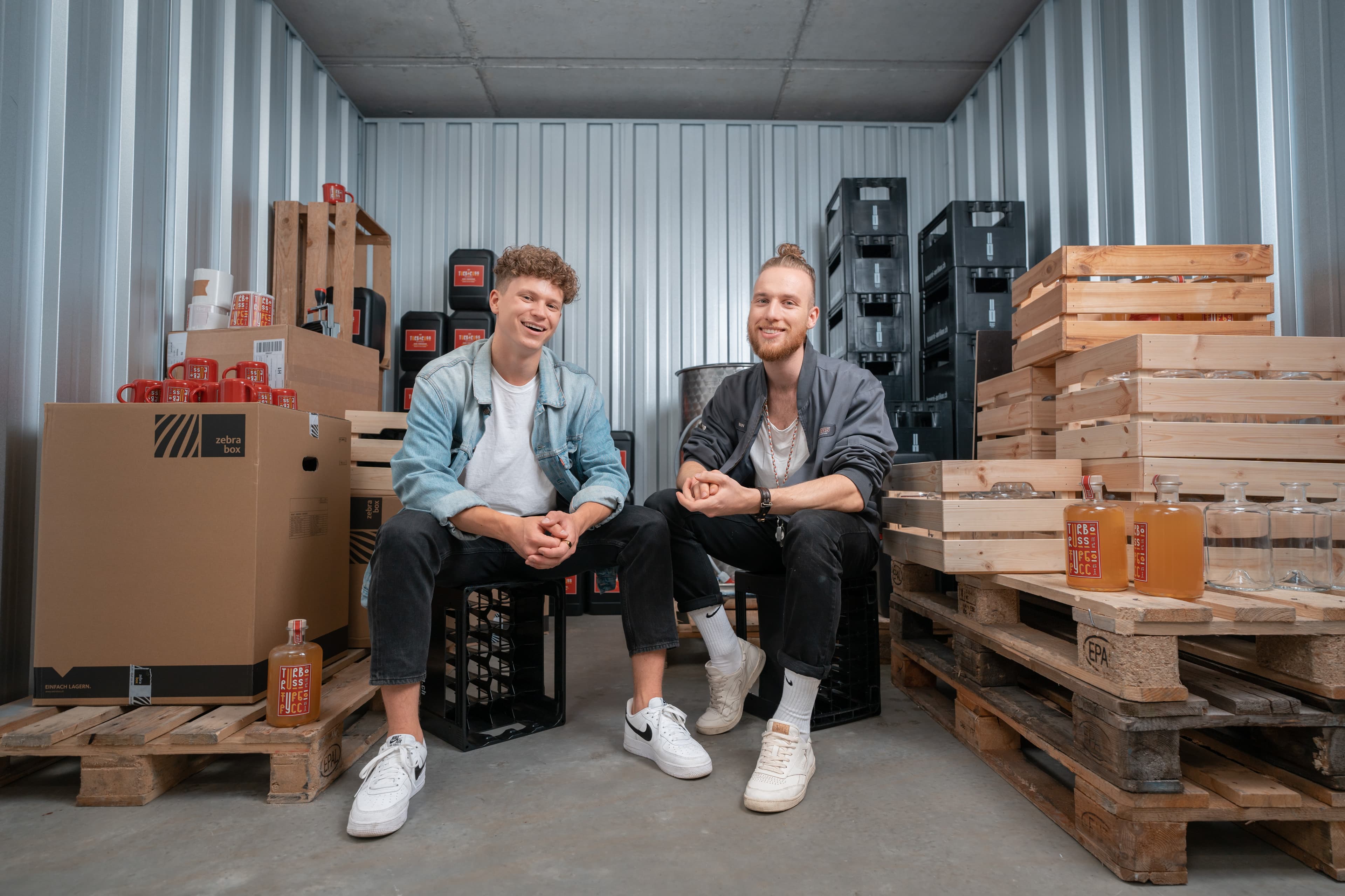 Zwei junge Startup Gründer einer Brauerei sitzen im Lagerraum von Zebrabox und schauen fröhlich in die Kamera.